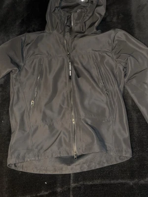 Svart vindjacka från C.P. Company - Snygg svart vindjacka från C.P. Company med huva och dragkedja framtill. Jackan har två snedställda fickor med dragkedja och är tillverkad i återvunnen nylon med Chrome-R teknologi. Perfekt för dig som gillar stilrena och funktionella plagg. Storlek S men passar även M