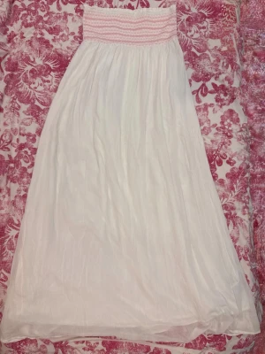 Vit långklänning med smock - Söt vit långklänning med bred smock upptill i rosa och vitt. Klänningen har en luftig och flowy passform, perfekt för varma dagar. Tillverkad i ett lätt tyg som ger en avslappnad känsla.
