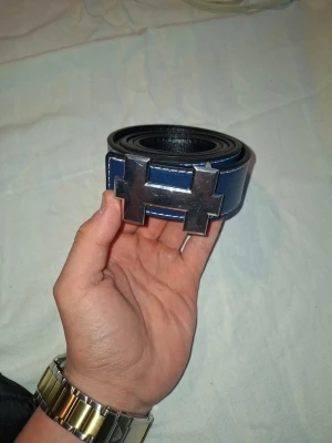 Blått skärp med H-spänne - Snyggt blått bälte med ett stort, metalliskt H-format spänne. Bältet har vita sömmar längs kanterna och är gjort i skinn. Perfekt för att lyfta vilken outfit som helst med en cool detalj. Fås med hålgörare, helt nytt skick!