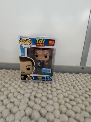 Funko Pop! Sid Toy Story  - Säljer en Funko Pop! vinylfigur av Sid från Toy Story, nummer 1598. Figurerna är detaljerade med Sid i svart t-shirt med dödskalle och leksaker i händerna. Förpackningen har Toy Story 30-årsjubileumslogga och färgglad design.