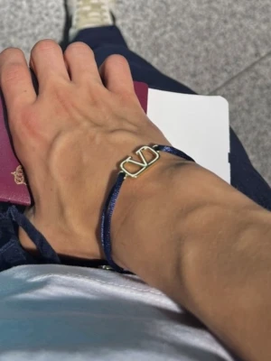 Valentino armband med blått band - Snyggt armband från Valentino med justerbart blått tygband och guldfärgat V-logo i metall. Perfekt accessoar för att lyfta din stil med en lyxig touch. Enkelt att ta på och av tack vare knytningen.