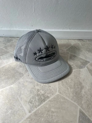 Grå meshkeps från Ciele Athletics - Snygg grå keps från Ciele Athletics med meshpaneler för extra ventilation. Framtill finns broderade svarta stjärnor och logga, samt justerbar snapback baktill. Perfekt för dig som vill ha en sportig och trendig look.