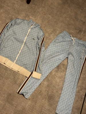 Tracksuit från Lacoste - Snyggt blått loungewear-set från Lacoste med diskret mönster och vita samt bruna detaljer. Setet består av ziptröja och byxor med dragsko i midjan. Båda plaggen har Lacoste-loggan broderad och vita muddar. Perfekt för sommaren.