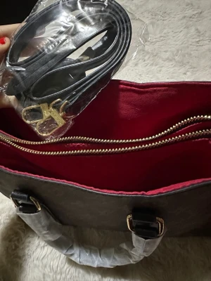 Fint handväska - Snygg handväska i klassiskt brunt monogrammönster med svarta detaljer och guldfärgade metalldelar. Insidan är fodrad i rött tyg och väskan har dubbla dragkedjor. Avtagbar axelrem i svart medföljer.