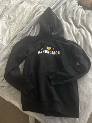 Svart hoodie BANANCIAGA från Jackie - Svart hoodie med huva och dragsnören med BANANCIAGA. Tryck med vit text och en gul banan på bröstet. Klassisk känguruficka framtill och ribbade muddar. Perfekt för dig som gillar streetwear och vill sticka ut med humoristisk stil. Normal i storleken. Ord pris 1195:-