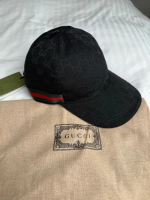 Svart keps från Gucci  - Snygg svart keps från Gucci med diskret GG-monogram över hela kepsen och klassiska gröna och röda ränder på sidan. Kepsen har böjd skärm och justerbar rem bak. Tillverkad i canvasmaterial och levereras med originalpåse.