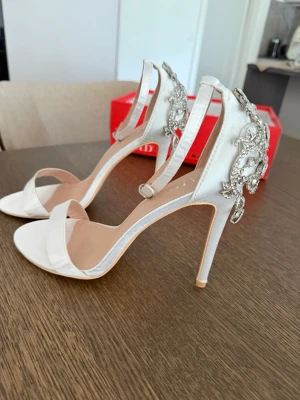 Vita pumps med strass från RAID - Säljer ett par vita pumps från RAID med öppen tå, smal rem över foten och hög klack. Baksidan pryds av stora, glittrande strassdetaljer som ger extra wow-känsla. Skorna har en elegant siluett och justerbar ankelrem. Perfekta för dig som vill sticka ut.