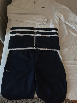 Lacoste vit och marinblå träningsdress - Snygg träningsdress från Lacoste i vitt och marinblått. Jackan har dragkedja, randiga detaljer och diskret Lacoste-logga på bröstet samt stor logga på ryggen. Byxorna är marinblå med elastisk midja och klassisk krokodil-logga. Perfekt för sport eller chill. Priset kan sänkas lite vid snabbt köp!