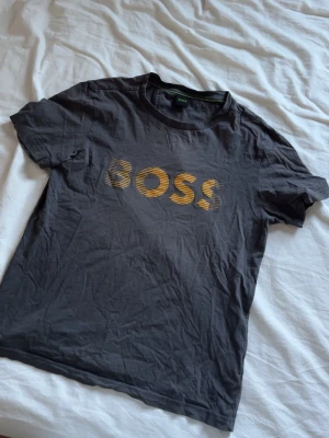 Svart BOSS t-shirt med gulddetalj - Snygg svart t-shirt från BOSS med guldfärgad logga framtill. Klassisk rund hals och korta ärmar. Tillverkad i mjuk bomull som känns skön mot huden. Perfekt för dig som gillar stilrena plagg med en touch av lyx. Xs men passar som s