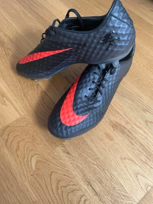 Nike svarta fotbollsskor med röd swoosh - Säljer ett par svarta Nike fotbollsskor med stor röd swoosh på sidan. Skorna har snörning och mönstrad yta för bättre bollkontroll. Sulan har fasta dobbar, perfekt för spel på gräs. Snygg och sportig design som sticker ut på planen.