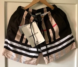 Burberry badbyxor med grafiskt mönster - Snygga badbyxor från Burberry med grafiskt mönster i svart, beige och vitt samt en diskret röd detalj. Modellen har elastisk midja med snörning och är tillverkad i lätt syntetmaterial som torkar snabbt. Perfekta för strand eller pool.