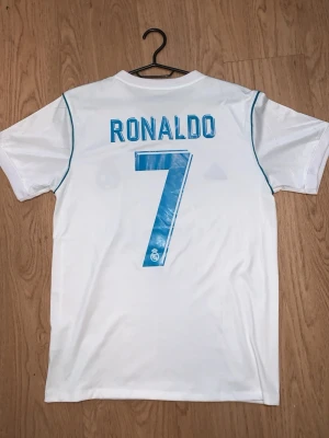 Real Madrid Ronaldo matchtröja - Säljer en vit Real Madrid matchtröja från Adidas med Ronaldo och nummer 7 på ryggen. Tröjan har blå detaljer, klubbmärke och Fly Emirates-tryck på bröstet samt klassiska Adidas-ränder på axlarna. Perfekt för fotbollsfans och samlare.