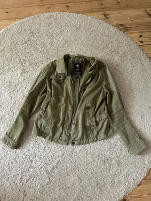 Olivgrön vindjacka - Snygg olivgrön vindjacka från G-Star RAW med dragkedja och tryckknappar framtill. Jackan har två fickor med lock och knappdetalj och en cool look.