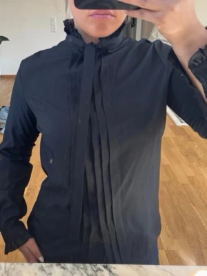 Svart blus med volangkrage och knytband - Stilren svart blus med lång ärm, volangdetaljer vid krage och ärmslut samt knytband i halsen. Framsidan har dekorativa veck som ger en elegant touch. Perfekt att matcha med både jeans och kjol för en trendig look.