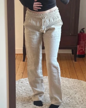 Beige linnebyxor - Säljer ett par beige linnebyxor med avslappnad passform och bred resår i midjan. Byxorna har en grov dragsko och stora fickor framtill. Perfekta för en chill och stilren look. Materialet är luftigt linne som känns svalt och skönt.