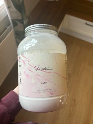 Protein Slim hallon & vit choklad - Stor burk måltidsersättning med smak av hallon och vit choklad från Protein. Pulver i en genomskinlig plastburk med silverfärgat lock och ljus etikett med rosa detaljer. Perfekt för smoothies eller shakes. Den inne håller 1,2 kg och har använt en gång så den är ny! Säljer så jag inte använder dom så dom står bara, bättre dom kommer till användning hos en annan!