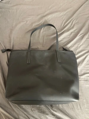 Svart shoppingväska i konstläder - Stilren svart väsk i konstläder med två långa handtag och dragkedja upptill. Väskan har en rymlig insida med innerfack och enkel, modern design som passar till det mesta. Perfekt för dig som vill ha plats för allt du behöver under dagen. Lite skitig på utsidan!