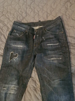 Mörkblå slitna jeans med hål - Säljer ett par mörkblå jeans med slitningar och ett större hål på vänster lår. Jeansen har klassisk femficksmodell, raka ben och detaljer i form av slitna partier och tvättade effekter. Perfekta för en avslappnad och trendig look.