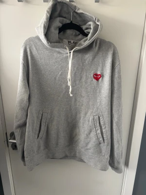 Grå hoodie från Comme des Garçons Play - Säljer en grå hoodie från Comme des Garçons Play med det ikoniska röda hjärtat med ögon broderat på bröstet. Hoodien har huva med vit dragsko och praktiska fickor framtill. Materialet är mjukt och bekvämt, perfekt för en chill look. Det står att tröjan är XL men den är betydligt mindre!