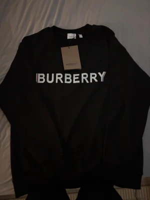 Svart sweatshirt från Burberry - Snygg svart sweatshirt från Burberry med stor vit logga över bröstet. Klassisk rund halsringning och ribbade muddar vid ärmslut och nederkant. Tillverkad i mjukt bomullsmaterial för skön känsla. Perfekt för dig som gillar stilrena designer och streetwear-vibe.