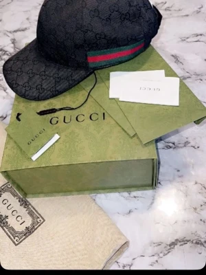 Svart Gucci keps - Säljer en svart keps från Gucci med klassiskt GG-monogram över hela kepsen. På sidan finns en grön och röd rand som signaturdetalj. Kepsen är i tygmaterial och har böjd skärm. Kommer med originalkartong, dustbag och äkthetskort. Använd ett par gånger och köpt från Guccis hemsida 