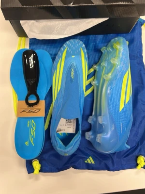 Adidas F50 blåa fotbollsskor - Säljer ett par Adidas F50 fotbollsskor i blått med gula detaljer och klassiska stripes. Skorna har en strömlinjeformad design, tillverkade i lätt syntetmaterial för maximal fart på planen. Levereras med extra innersulor och skopåse. Perfekta för dig som vill sticka ut!