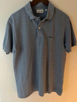 Blå pikétröja från Lacoste - Klassisk blå pikétröja från Lacoste med korta ärmar och den ikoniska krokodilloggan på bröstet. Tröjan har krage och knäppning med två knappar framtill. Perfekt för en clean och stilren look.