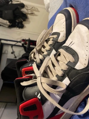 Puma vita och svarta high sneakers - Snygga sneakers från Puma i vitt, svart och rött. Klassisk high-top modell med kraftig sula och beige snörning. Skorna har Pumas ikoniska logga på sidan och röd detalj vid hälen. Perfekta för dig som gillar retrostil och streetwear.