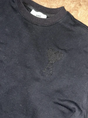Svart sweatshirt från Ami Paris - Svart sweatshirt från Ami Paris med broderad ton-i-ton logga i form av ett hjärta med bokstaven A på bröstet. Tröjan har rund halsringning, långa ärmar och ribbade muddar. Perfekt för en clean och stilren look.