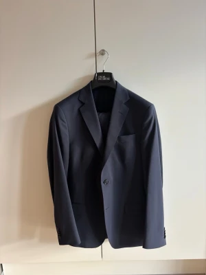 Oscar Jacobson Edmund Navy Suit - Säljer min Oscar Jacobson-kostym som är i absolut nyskick, endast använd vid två tillfällen. Både jacka och byxa är helt oförändrade och utan skräddarförändringar (bortsett från att bröstfickan sprättats upp).  Modellen är Edmund (enkelknäppt), som är en smalare passform. Både jacka och byxa är i storlek 48. Färgen är mörkblå. Kostymen är köpt för 5 998 SEK på Oscar Jacobsons hemsida. För exakta mått hänvisas till deras storleksguide på deras hemsida, då kostymen är helt ojusterad.