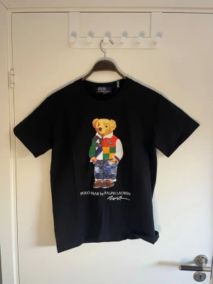 Svart Polo Bear t-shirt från Ralph Lauren - Säljer en svart t-shirt från Ralph Lauren med den ikoniska Polo Bear-trycket på framsidan. T-shirten har rund hals och korta ärmar. Materialet är mjuk bomull och passformen är normal. Perfekt för dig som gillar klassiska streetwear-vibbar.