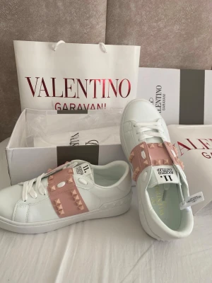 Valentino Garavani vita sneakers - Säljer ett par vita sneakers från Valentino Garavani med breda ljusrosa band över vristen dekorerade med ikoniska nitar. Skorna har klassisk låg profil, vita snören och detaljer i läder. Perfekta för dig som vill ha en lyxig och trendig look.