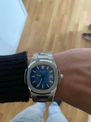 Silvrig blå klocka - Säljer en stilren klocka med blå urtavla och silverfärgat armband i rostfritt stål. Klockan har ett klassiskt, sportigt utseende med datumfönster och tydliga indexmarkeringar. Perfekt accessoar för dig som gillar exklusiv design.