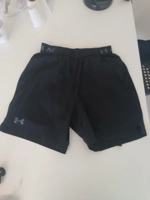 Svarta träningsshorts Under Armour - Säljer ett par svarta träningsshorts från Under Armour med elastisk midja och logga på benet. Shortsen är tillverkade i ett lätt och snabbtorkande material, perfekta för gymmet eller löpning. Snygg och sportig design med diskreta detaljer.