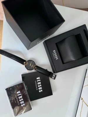 Svart klocka från Versus Versace - Snygg svart klocka från Versus Versace med gulddetaljer och stilren urtavla. Klockan har ett svart läderband och coola dekorativa element på boetten. Kommer i originalbox med tillhörande manual och certifikat.