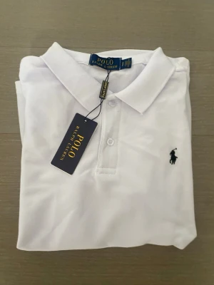 Vit pikétröja från Ralph Lauren - Klassisk vit pikétröja från Ralph Lauren med korta ärmar och krage. Tröjan har knappar framtill och den ikoniska broderade loggan i svart på bröstet. Perfekt till jeans eller shorts för en clean och stilren look.