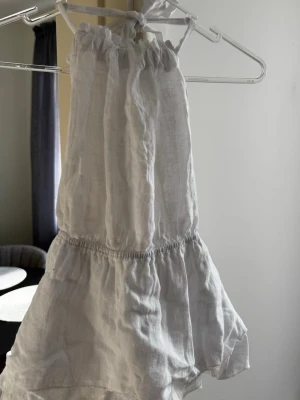 Vit linnetopp med volang från Designbysi  - Luftig och ljus vit linnetopp från DesignBySi med knyt i nacken och volangkant nedtill. Toppen är ärmlös och har öppen rygg, vilket ger en fräsch och somrig vibe. Perfekt att styla till jeans eller kjol för en trendig look. Aldrig använd, prislapp kvar.