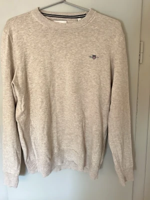 Beige tröja från Gant med brodyr - Klassisk beige tröja från Gant med rund halsringning och diskret broderad logga på bröstet. Tröjan har ribbade muddar vid ärmslut och nederkant. Perfekt för en clean och stilren look.