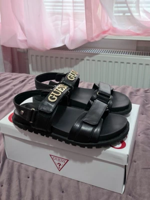 Guess Sandaler  - Säljer ett par supersnygga och stilrena sandaler från Guess i svart med gulddetaljer. De är köpta förra året i Stockholm  och användes endast under förra sommaren, så de är fortfarande i jättefint skick! Kartongen följer med  