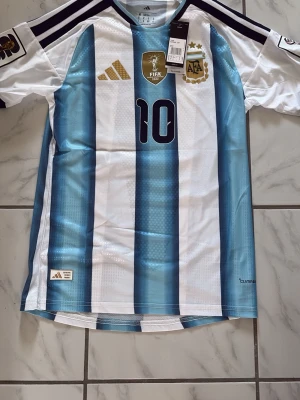 Argentina Messi matchtröja Adidas - Säljer en officiell Argentina fotbollströja från Adidas med Messi och nummer 10 på ryggen. Tröjan har klassiska blå och vita ränder, svarta detaljer och AFA-emblemet på bröstet. Perfekt för dig som älskar fotboll och vill stötta Argentina.