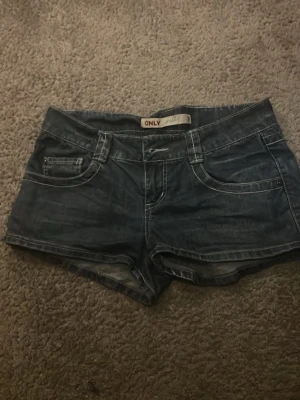 Mörkblå jeansshorts från Only - Snygga mörkblå jeansshorts från Only med klassisk femficksdesign och kontrastsömmar. Shortsen har bälteshällor, knappgylf och coola detaljer på bakfickorna. Perfekta för varma dagar och enkel att matcha med olika toppar.🌸