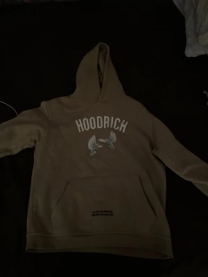 Beige hoodie från Hoodrich med tryck - Säljer en beige hoodie från Hoodrich med stor huva och känguruficka. Framsidan har HOODRICH-tryck och två duvor, samt texten 'ALWAYS PROUD NEVER SATISFIED' på fickan. Baksidan har stort duvtryck och text i skrivstil. Perfekt för en streetwear-look.