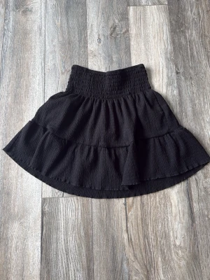 Svart volangkjol från Gina Tricot - Supersnygg svart kjol från Gina Tricot med bred smock i midjan och dubbla volanger. Kjolen har en kort och flowy modell som passar perfekt till alla dina outfits. Tillverkad i ett mjukt och lätt material som ger en skön känsla.