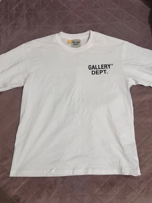 Vit t-shirt från Gallery Dept. - Säljer en vit t-shirt från Gallery Dept. med svart tryck fram och stort tryck på ryggen där det står 'GALLERY DEPT. HOLLYWOOD, CA.'. Klassisk passform och rund hals. Perfekt för dig som gillar streetwear och enkel stil.