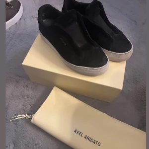 Svarta sneakers från Axel Arigato  - Säljer ett par svarta sneakers från Axel Arigato med vit sula och minimalistisk design. Skorna har låg profil och är utan snörning, vilket ger en clean look. Perfekta för dig som gillar stilrena och moderna skor. Skorna är i storlek 36 men har töjts ut sig lite så att skulle kunna passa 37 väl. Med kommer med box och dust bag.