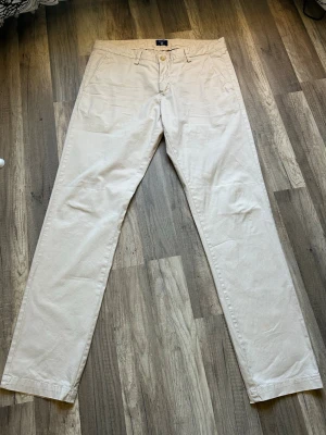 Beige chinos från Gant - Snygga beige chinos från Gant med klassisk rak passform. Byxorna har två sidofickor fram och två bakfickor med knapp. Tillverkade i mjukt bomullstyg som känns skönt mot huden. Perfekta för en clean och stilren look.