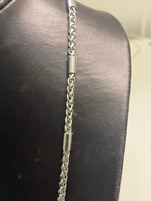 Silverfärgat kedjehalsband i stål - Snyggt halsband i silverfärgat stål med flätad kedja och cylinderformade detaljer längs hela halsbandet. Perfekt accessoar för att lyfta din outfit med en stilren och modern touch. Passar både till t-shirt och skjorta.