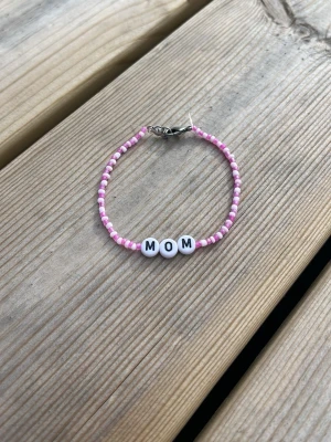 Pärlarmband med texten MOM - Sött armband med små rosa och vita pärlor samt tre vita pärlor i mitten med bokstäverna M O M. Armbandet har ett metallspänne och är perfekt för att visa uppskattning till mamma. Passar dig som gillar personliga accessoarer. Armbandet är 18 cm i längden.💓
