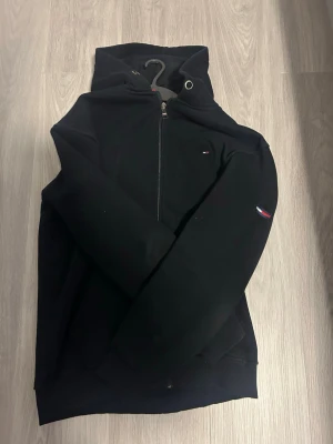 Svart hoodie från Tommy Hilfiger - Snygg svart hoodie från Tommy Hilfiger med halv dragkedja framtill och klassisk huva. Diskret Hilfiger-logga på bröstet och ärmen. Tillverkad i mjukt bomullsmaterial som känns skönt mot huden. Perfekt för en avslappnad och stilren look. Den har använts lite men är i väldigt bra skick utan några skador. Står att storleken är Xl men den sitter som M