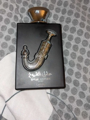 Lattafa Ishq Al Shuyukh Silver parfym - Lattafa Ishq Al Shuyukh Silver parfym i en svart, rektangulär flaska med en stor silverfärgad dekor och lock. Flaskan har en unik metallisk dekoration på framsidan och arabiska samt engelska texter. Perfekt för dig som gillar exklusiv design.